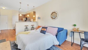 Un dormitorio moderno con cama, escritorio y silla. Se observa una zona de cocina al fondo.