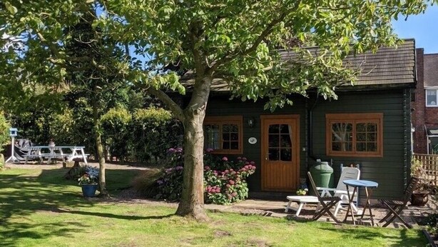 Une petite maison en bois verte, avec un grand arbre devant, un jardin fleuri et du mobilier d’extérieur.