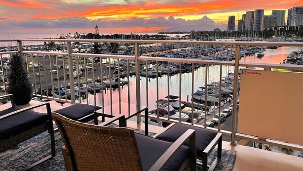 Een balkon met rieten meubilair, uitkijkend op een jachthaven met boten en een stadsachtergrond tijdens de zonsondergang.