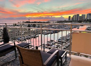 Varanda com mobiliário de vime, com vista para uma marina com barcos e o horizonte da cidade ao pôr do sol.