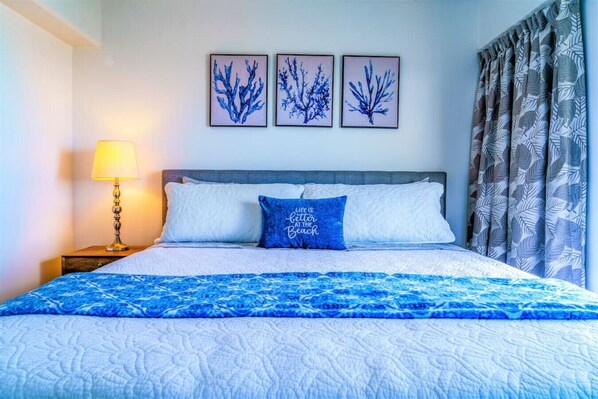 Una cama bien hecha con una colcha azul y blanca estampada, un cojín azul con una cita, y tres cuadros en la pared sobre ella.
