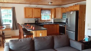 A compact kitchen with wooden cabinets, a stainless steel refrigerator, and a dining area with a long table and chairs. - Private quiet home close to Sunday River, Bethel village, and summer recreation  (Bethel)