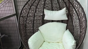 Un fauteuil suspendu en osier, doté de coussins, se trouve dans une pièce dont un mur est recouvert de carreaux et qui possède une fenêtre.
