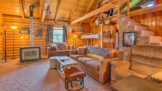 A log cabin interior with a stone staircase, wooden furniture, a fireplace, and a television. - Charming Montana Retreat w/ Mountain Views! (Checkerboard)