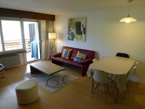 A living room with a red sofa, a coffee table, a dining table with chairs, a lamp, and a painting on the wall.