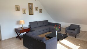 Ein modernes Wohnzimmer mit einem Ecksofa, zwei Sessel, einem Couchtisch und einem Beistelltisch mit Lampe. An der Wand befinden sich gerahmte Bilder.