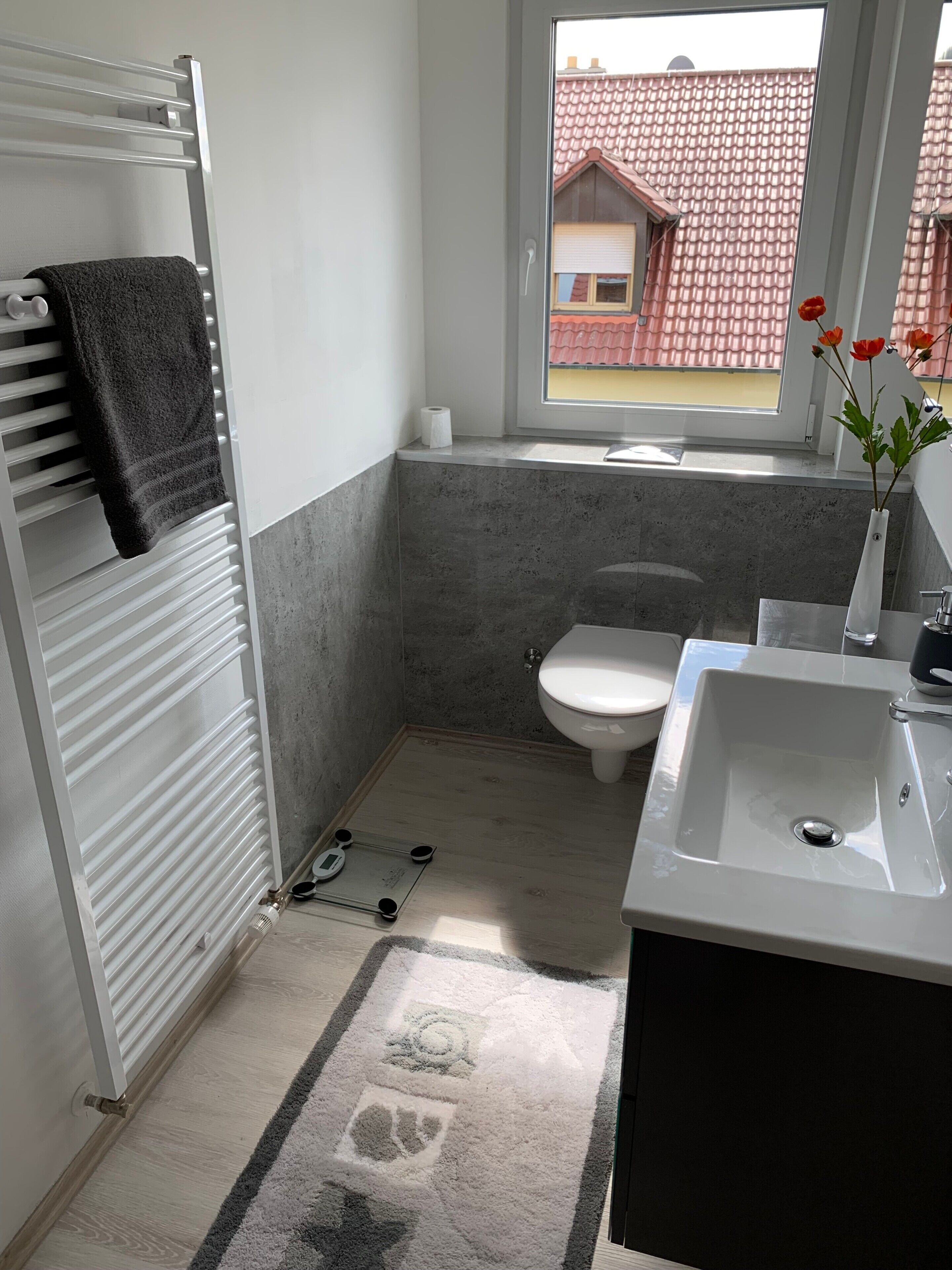 Un baño pequeño con un radiador blanco, un inodoro, un lavabo y una ventana con vistas a un edificio de techos rojos.