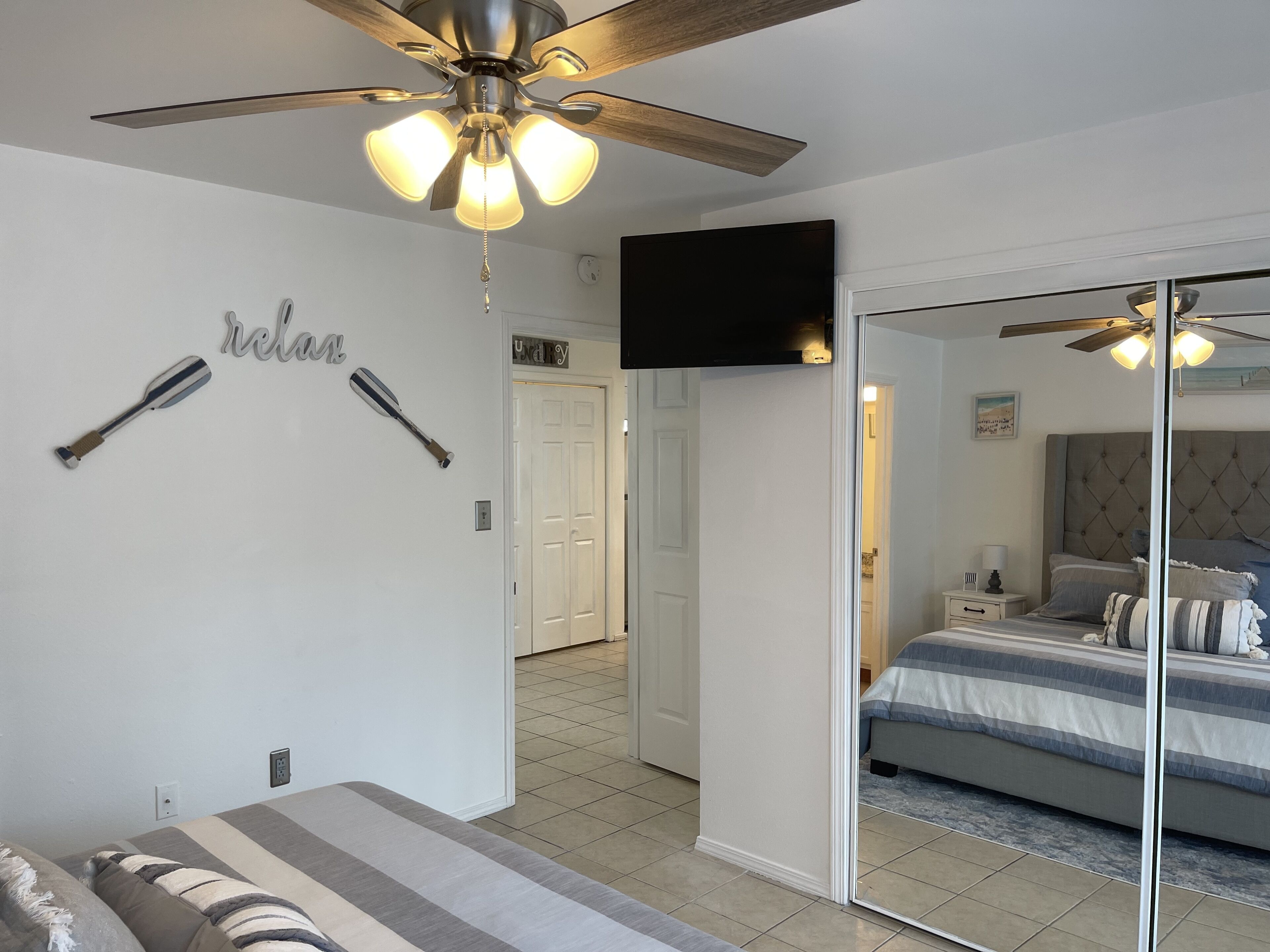 Une chambre à coucher avec un lit, un ventilateur de plafond, un téléviseur fixé au mur et un miroir qui reflète la pièce.