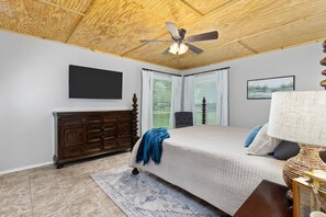 Ein Schlafzimmer mit Holzboden, einem Bett mit weißer Bettdecke, einer dunkelholzigen Kommode, einer blauen Tagesdecke, einem Deckenventilator und einem Fenster mit Jalousien.