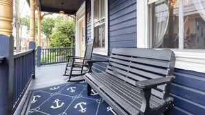 Une véranda avec un banc en bois et un fauteuil à bascule, un tapis décoratif orné de motifs d’ancres, et une vue sur la rue et la verdure.