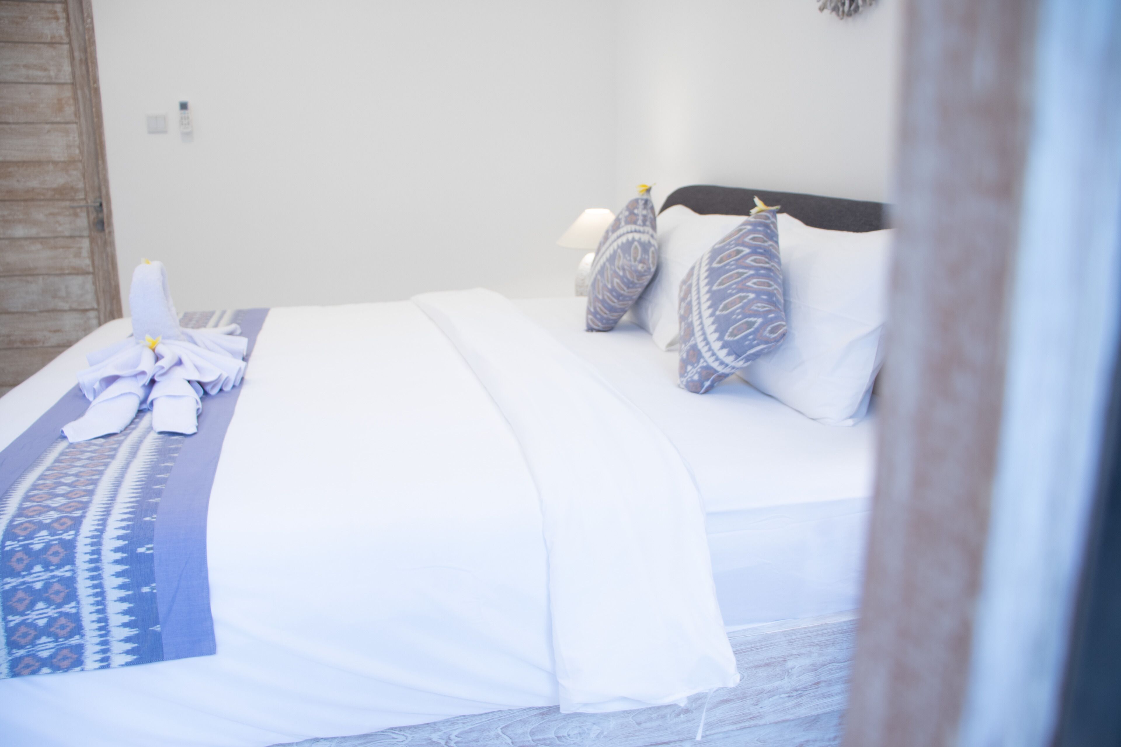 Ein ordentlich bezogenes Bett mit weißer Bettwäsche und gemusterten Kissen, ein Nachttisch mit einer Lampe und eine Holztür.