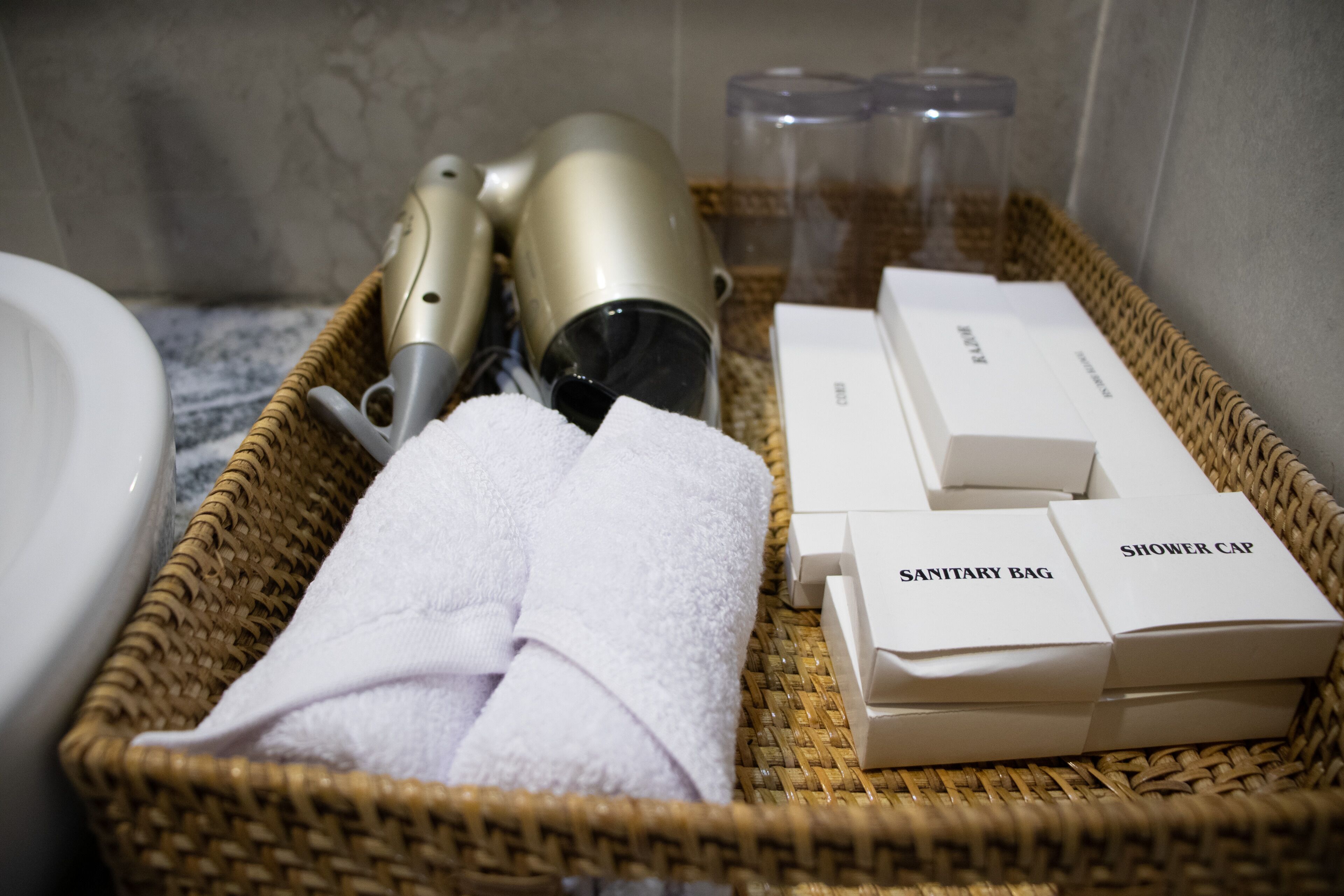Un panier en osier contenant une serviette blanche, un sèche-cheveux et deux boîtes étiquetées « SAC D’HYGIÈNE » et « CAPPE DE DOUCHE », le tout dans une salle de bain.