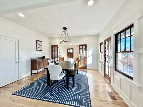A spacious dining area with a large wooden table, white chairs, and a blue patterned rug.