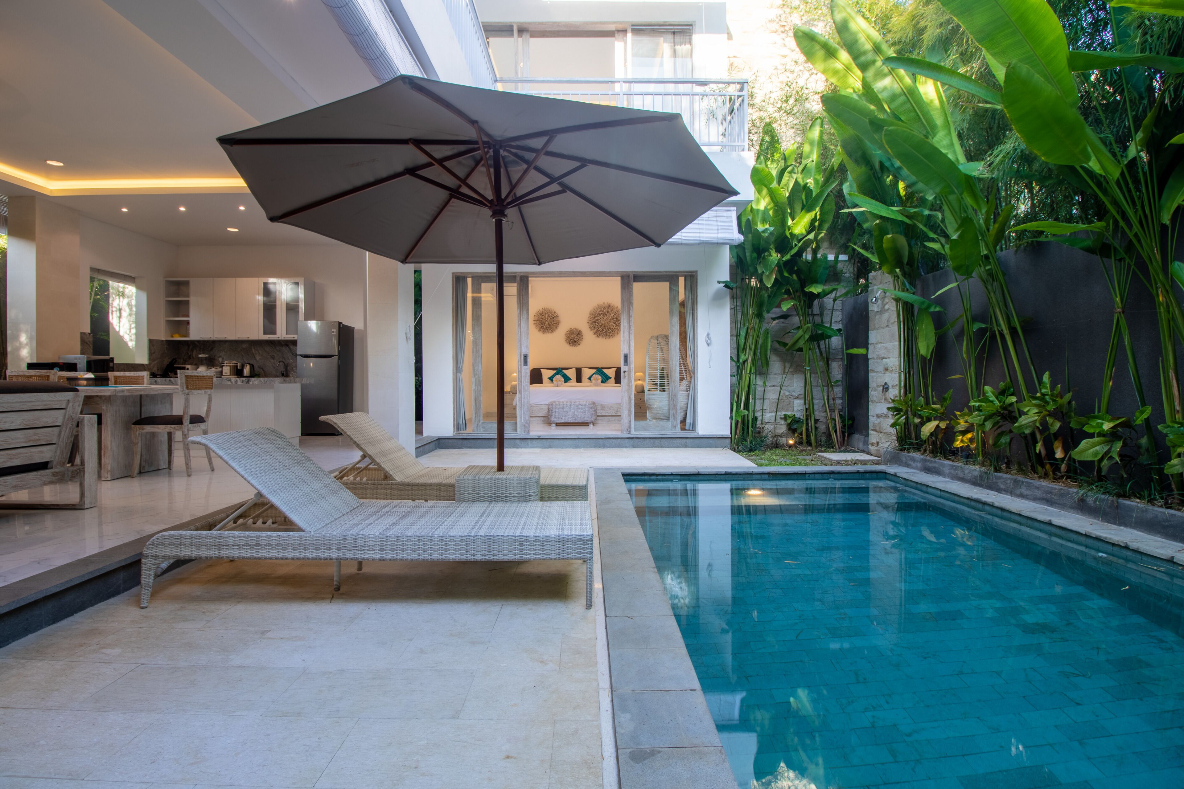 Un área de piscina moderna con sillones de descanso y una sombrilla, rodeada de exuberante vegetación y un espacio interior bien iluminado.