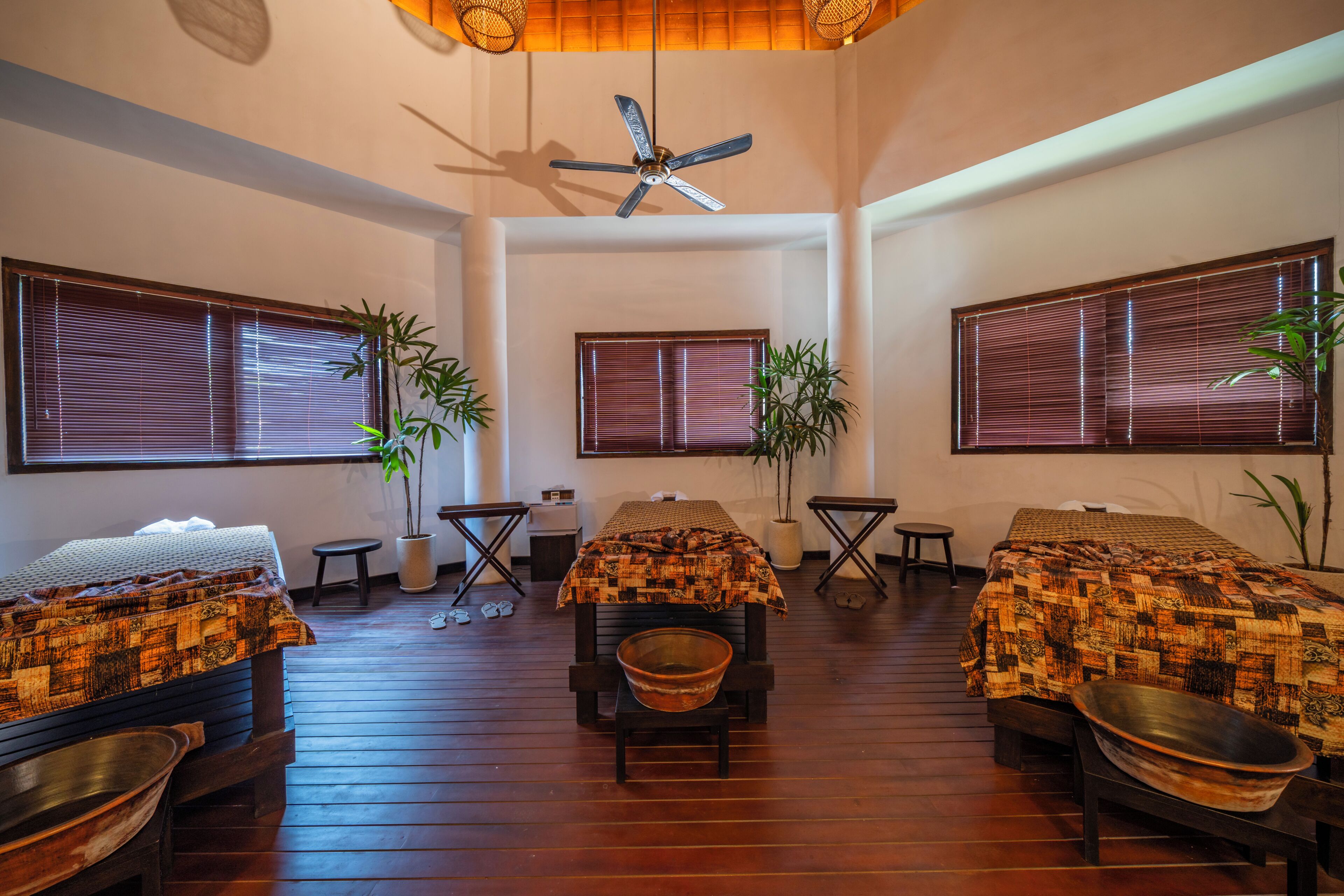 A room with a wooden floor, two beds with patterned blankets, a ceiling fan, and large windows with blinds.