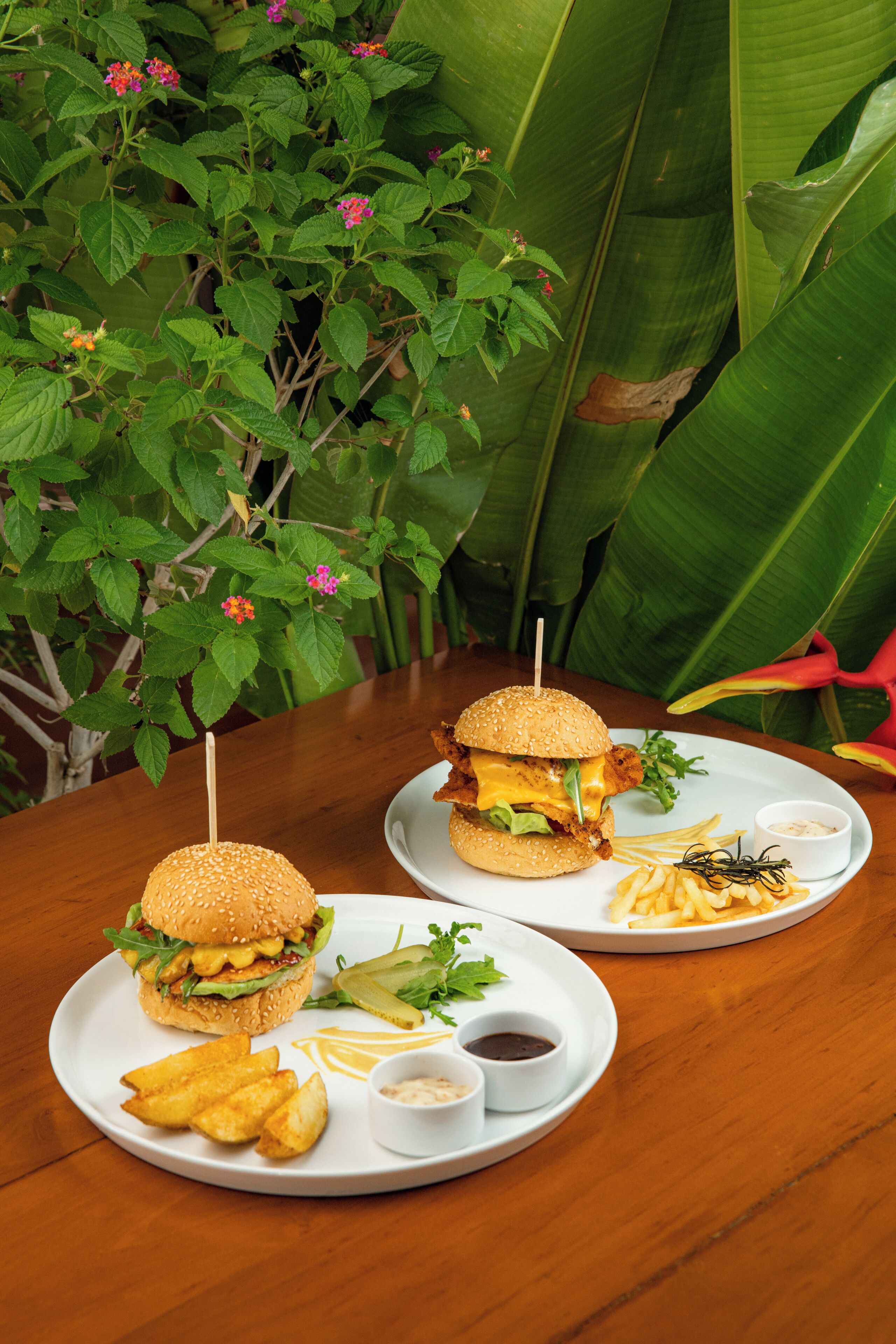 Two burgers with fries and condiments on plates, set on a wooden table with a tropical plant background.