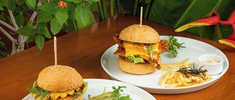 Two burgers with fries and condiments on plates, set on a wooden table with a tropical plant background.