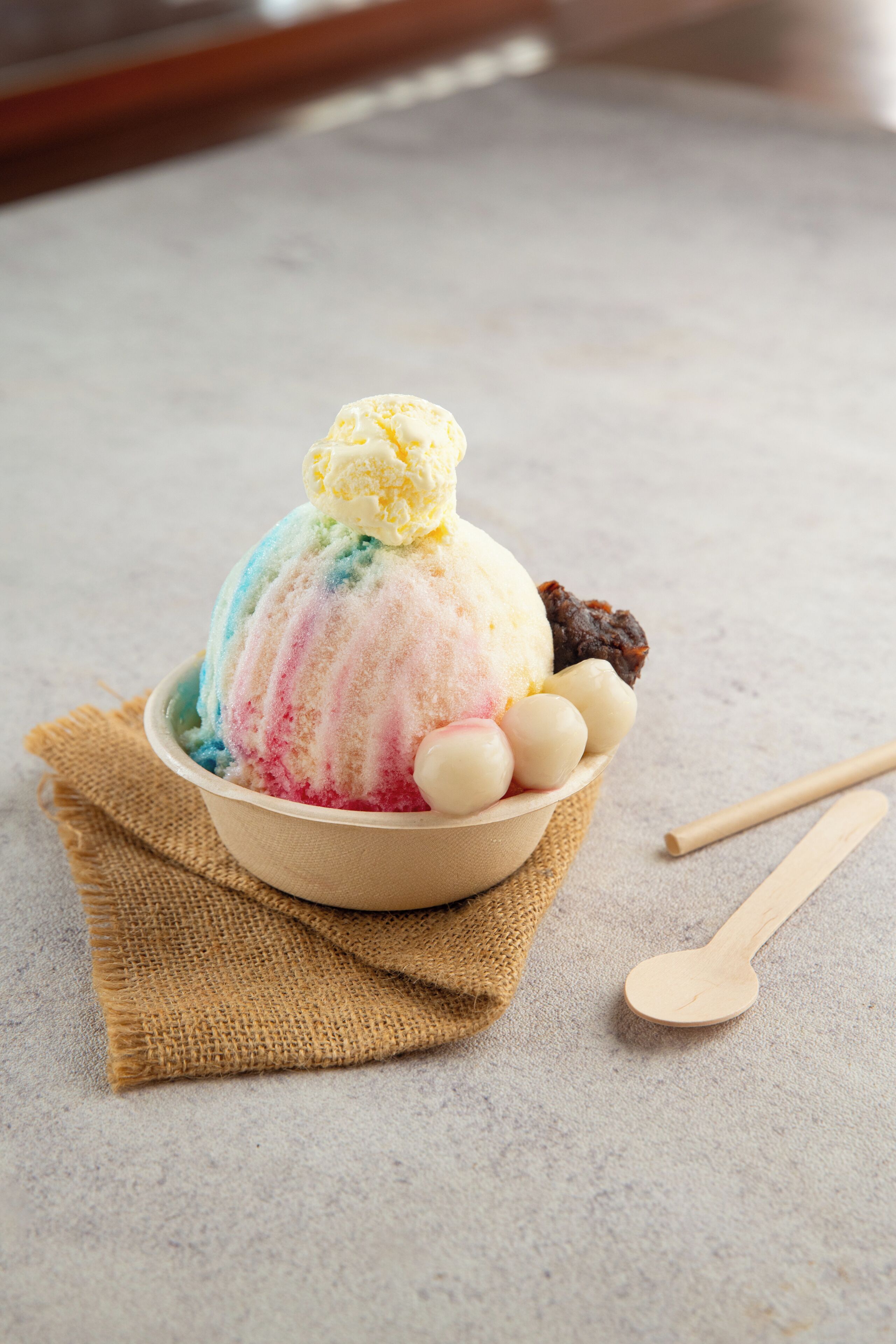 Eine Schale buntes Eis mit einer Kugel obenauf, begleitet von einem Holzlöffel und einem Stück Jutegewebe.