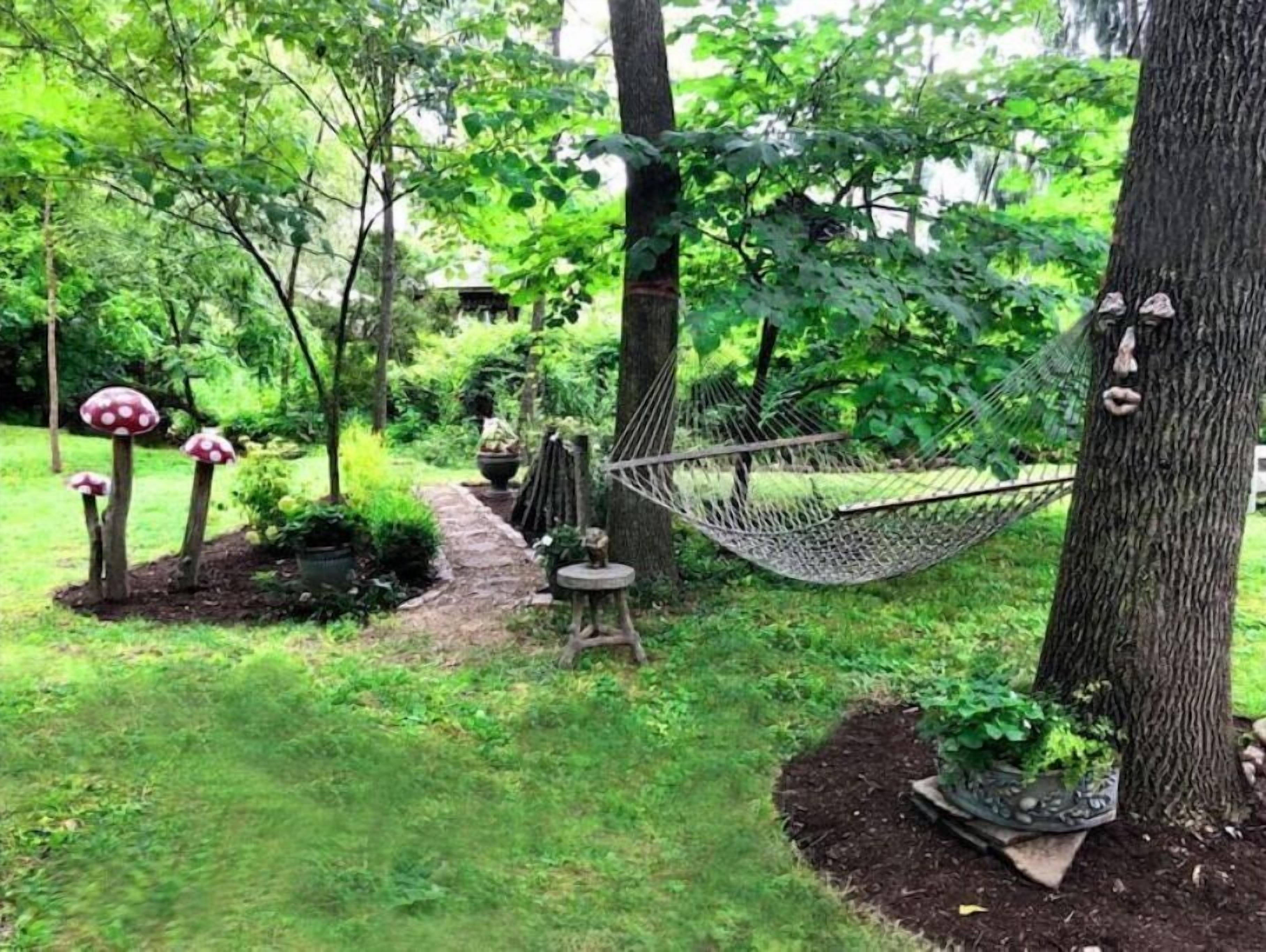 Eine Hängematte, die zwischen Bäumen in einem üppigen Garten gespannt ist, mit einem Steinweg und dekorativen Pilzskulpturen.