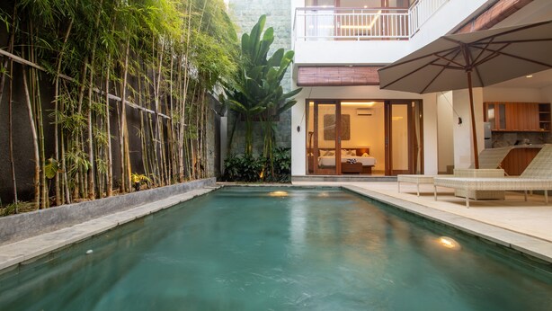 A swimming pool with a bamboo garden on one side and a modern house with a balcony and outdoor seating area on the other. - Sayang Sanur Villa VII (Denpasar)