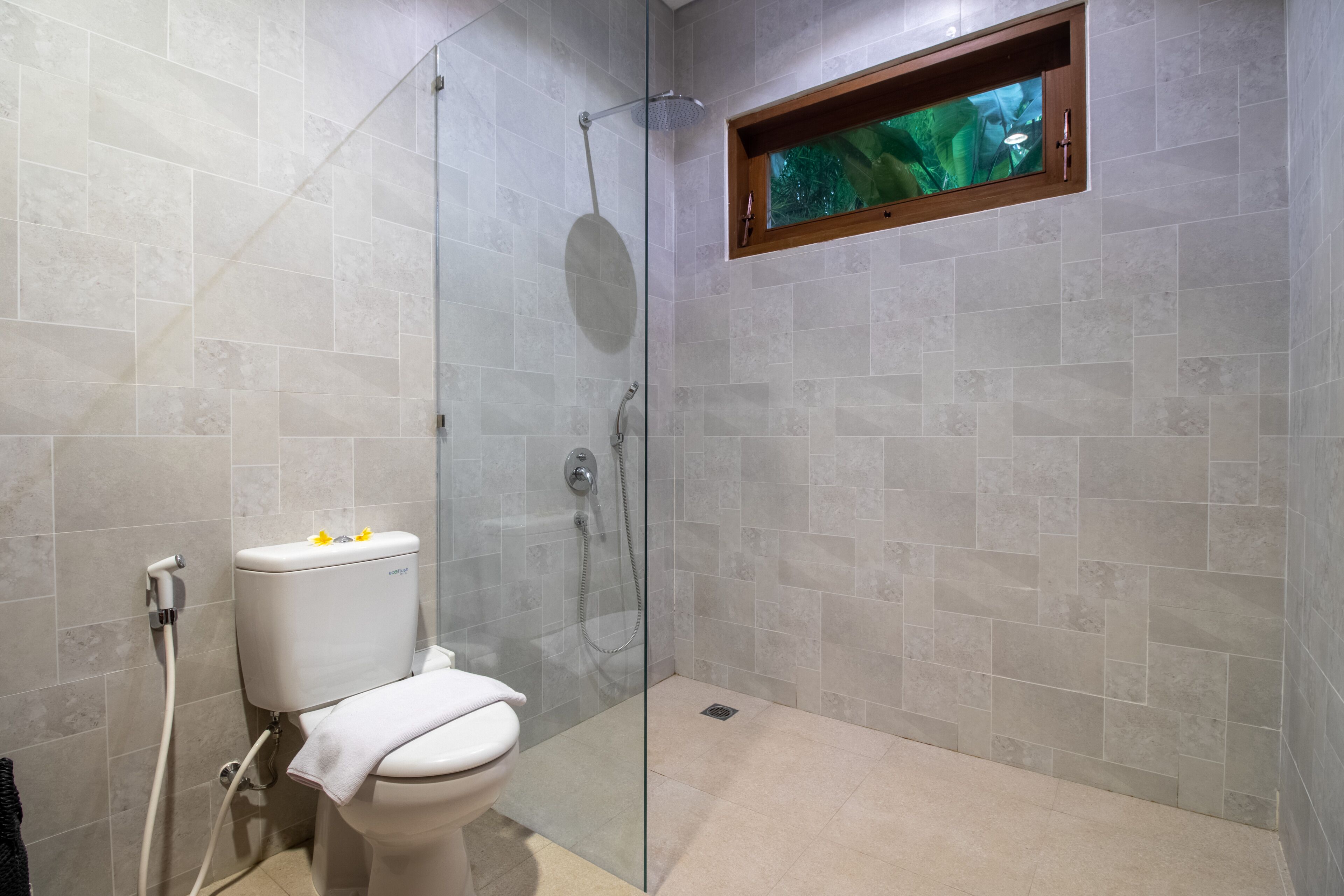 A modern bathroom with a glass-enclosed shower, a toilet, and a window with a view of greenery.
