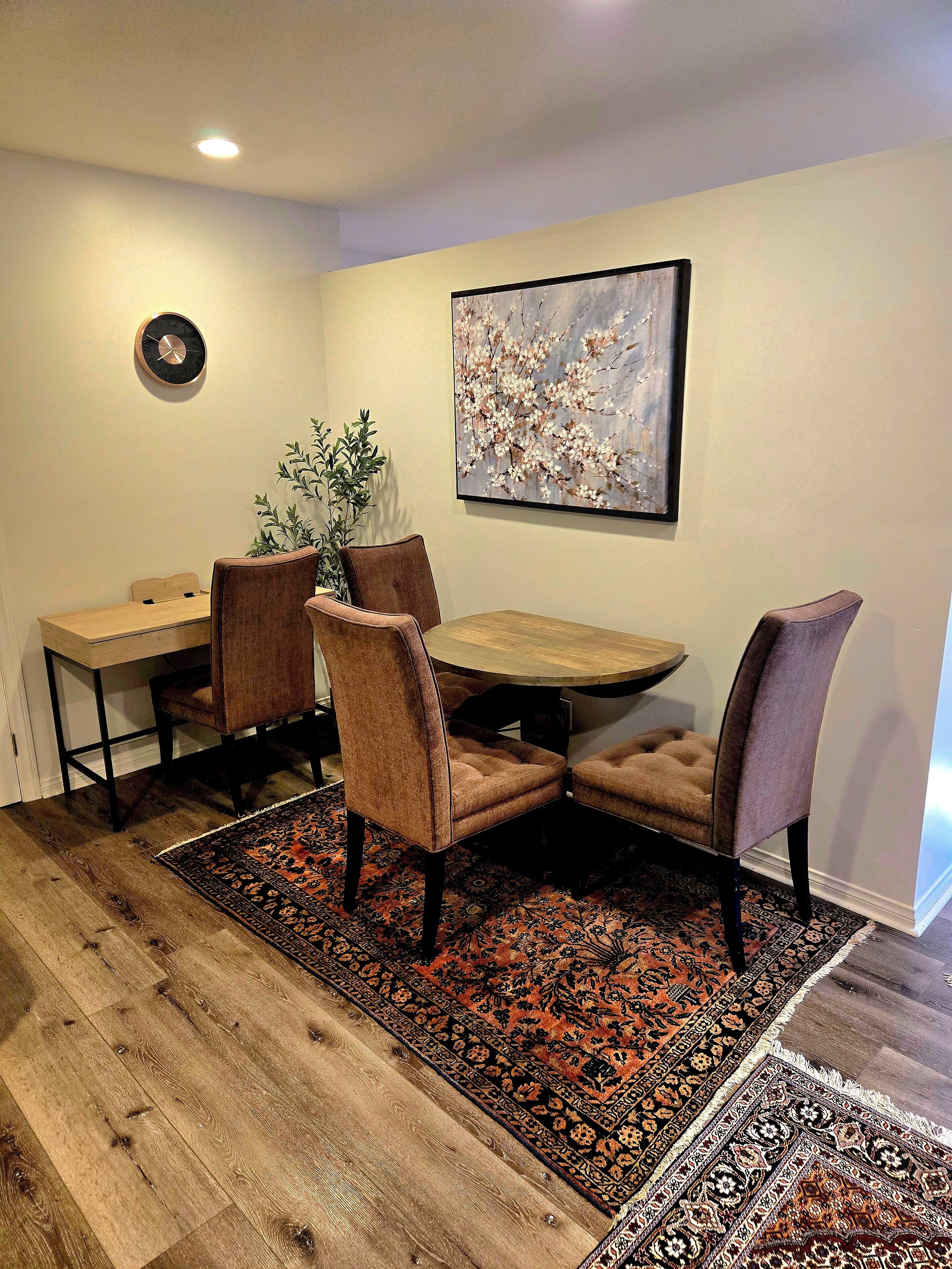 A dining area with a wooden table, four chairs, a patterned rug, and a wall-mounted clock.