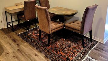 A dining area with a wooden table, four chairs, a patterned rug, and a wall-mounted clock.