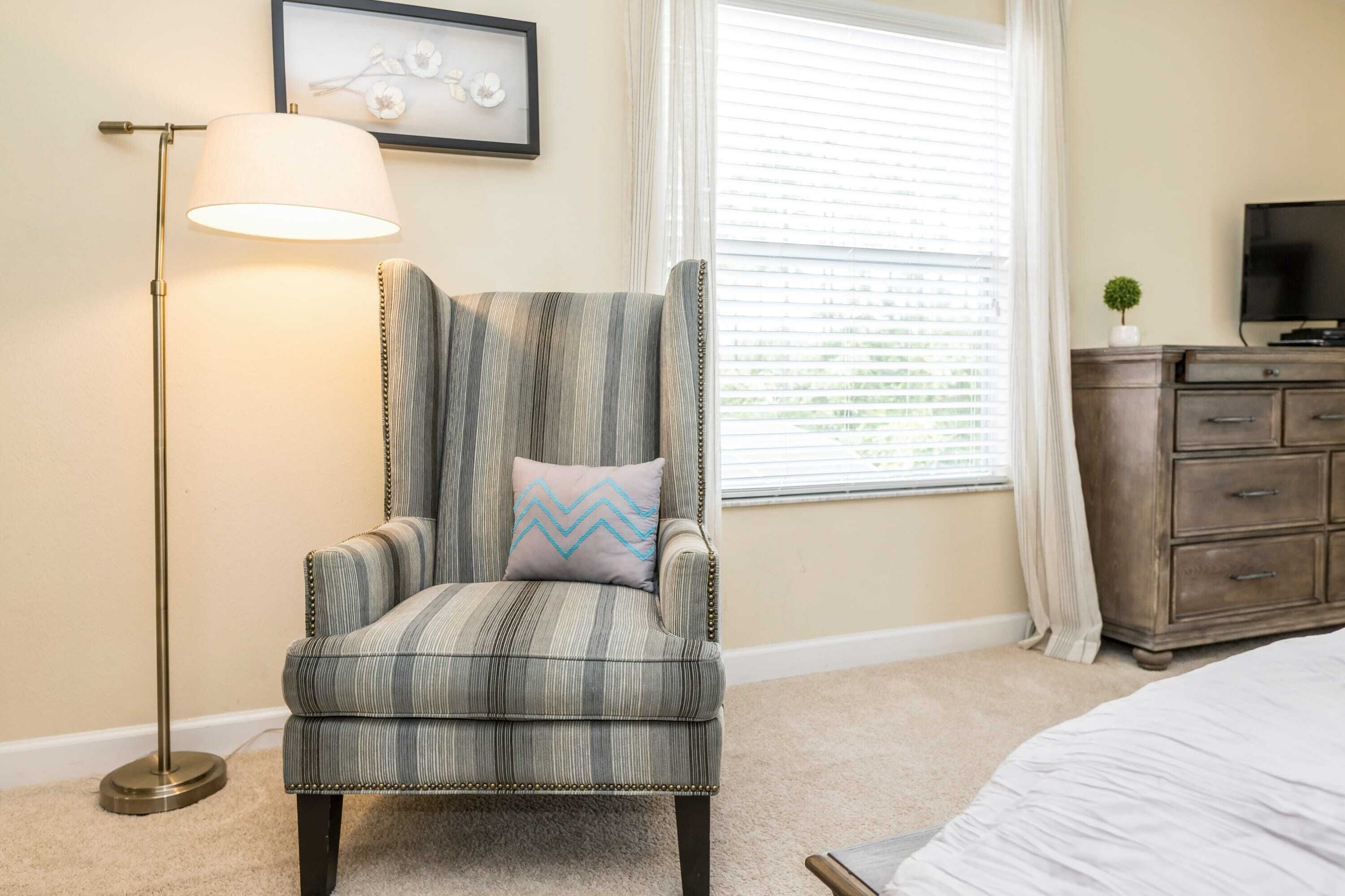 A room with a striped armchair, a floor lamp, a television on a wooden dresser, and a window with blinds.