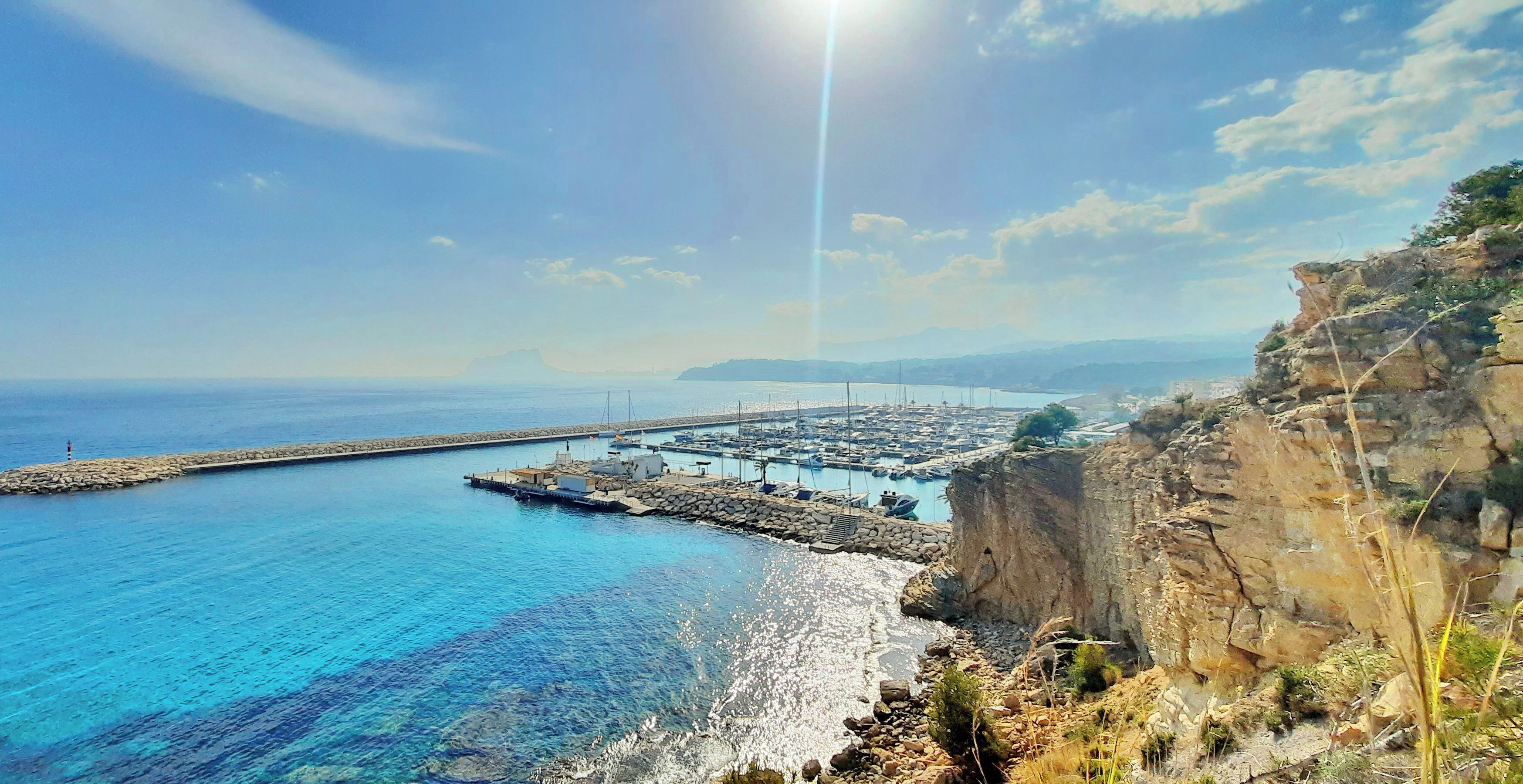 A coastal marina with boats docked, a rocky shoreline, and a clear sky.