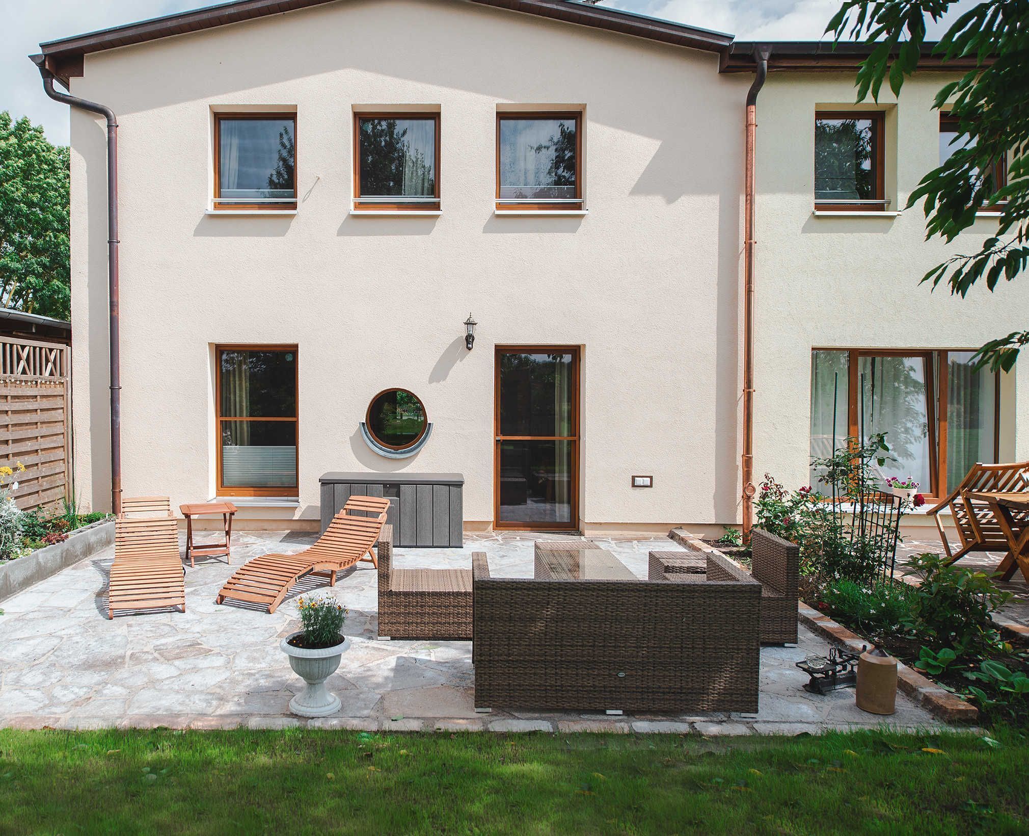 A two-story building with a patio area featuring outdoor furniture, a circular window, and a small garden.