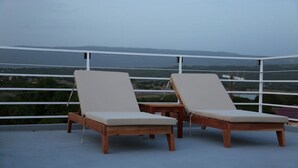 Two wooden lounge chairs with cushions on a balcony with a view of a landscape.