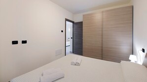 Habitación de hotel con cama, armario, mesita de noche con lámpara y puerta que da a otra habitación.