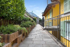 Un estrecho sendero de piedra bordeado por jardineras de madera con follaje, que conduce a un edificio amarillo con baranda metálica.