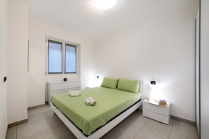A bedroom with a bed, bedside table, nightstand, window, and wall-mounted light.
