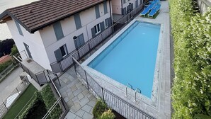 Een rechthoekig zwembad met een betegeld zonnedek, een gebouw met een rode pannendak en een hekwerk.