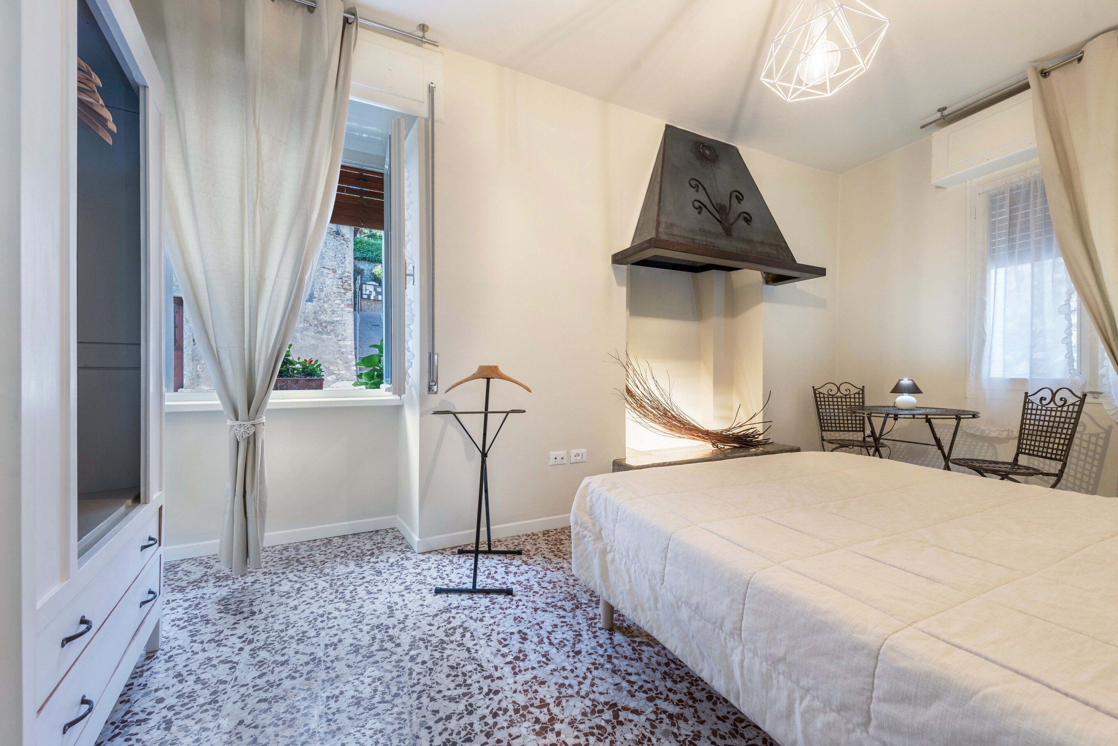 Een slaapkamer met een bed, een raam met gordijnen, een vloerlamp, een tafel met stoelen, en een decoratief object aan de muur.