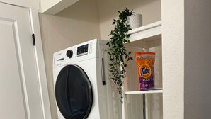 A laundry area with two stacked front-loading washing machines, a shelf with a plant and a Tide detergent box, and a white door.