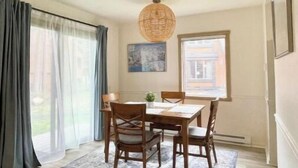A dining area with a wooden table and chairs, a woven light fixture, a painting on the wall, and a view through the window.