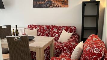 Een gezellige zithoek met een rode bloemenbank, een houten salontafel en een rieten fauteuil. Op de muur hangt een schilderij van een strandgezicht, en een vloerlamp staat op een plank.