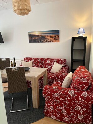 Ein gemütlicher Wohnbereich mit einem roten, floralen Sofa, einem Holztisch und einem Korbstuhl. Auf der Wand hängt ein Gemälde, das eine Strandansicht zeigt, und eine Stehlampe steht auf einem Regal.