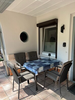 Une terrasse couverte avec une table et quatre chaises, une fenêtre et un luminaire fixé au mur.