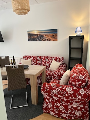 Un salón acogedor con un sofá floral rojo, una mesa de centro de madera y una silla de mimbre. Un cuadro de una playa cuelga en la pared, y una lámpara de pie está sobre una estantería.
