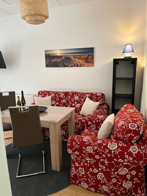 A cozy living area with a red floral sofa, a wooden coffee table, and a wicker chair. A painting of a beach scene hangs on the wall, and a floor lamp is on a shelf. - Apartment 5 - Karlshagen Apartments, Ute Deckert (Karlshagen)