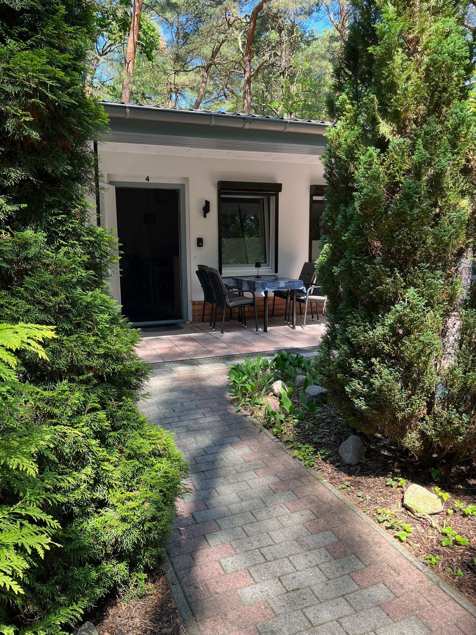 A modern building with a patio area featuring a table and chairs, surrounded by lush greenery and a brick pathway.