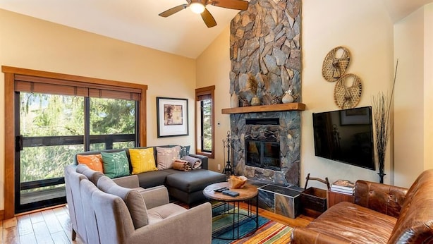 A modern living room with a stone fireplace, a ceiling fan, and a large window. - Ski In Whistler Village Townhome in Best Location Telemark (Whistler)