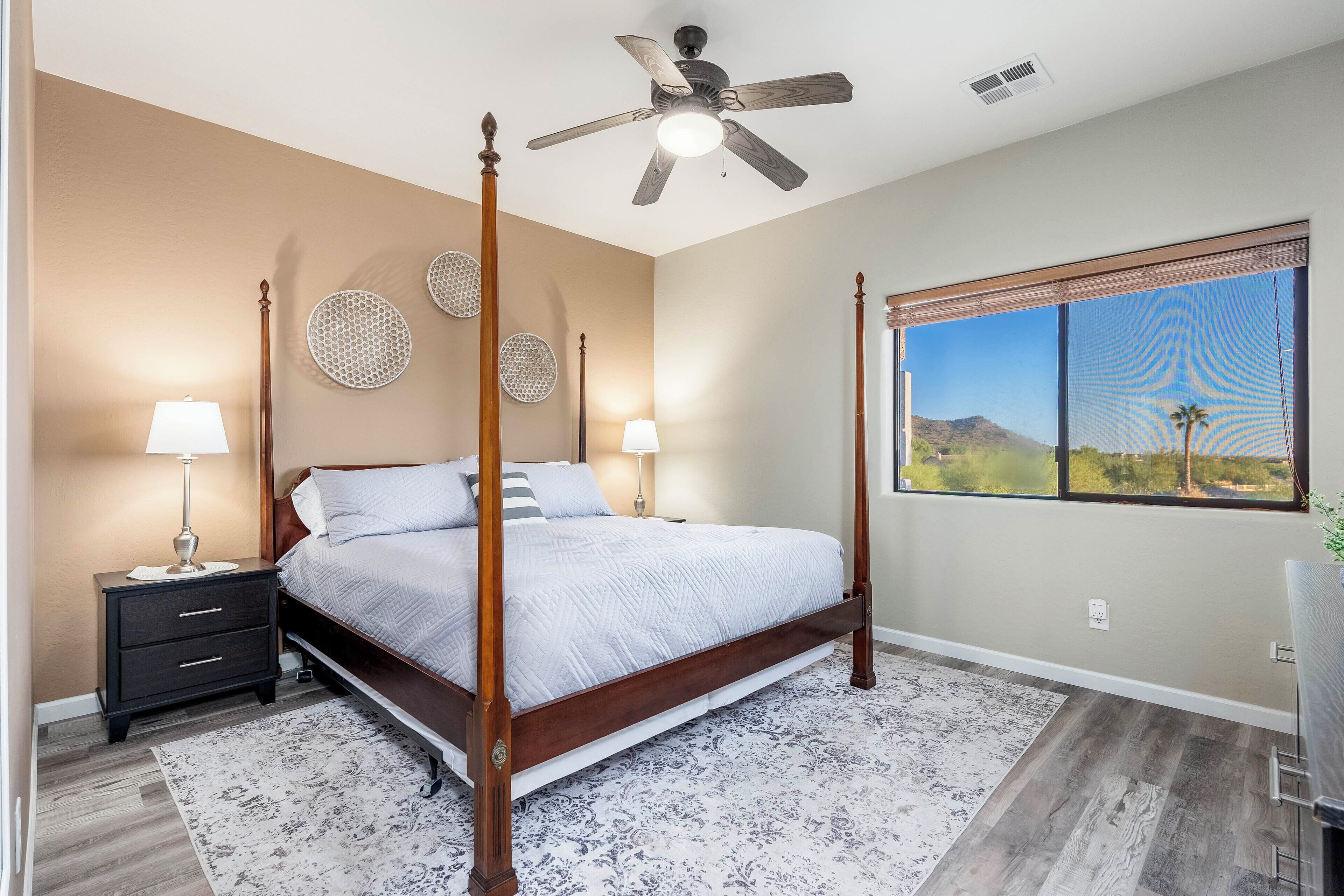 A bedroom with a four-poster bed, two bedside tables with lamps, a ceiling fan, and a large window with a view of a mountain and palm trees.