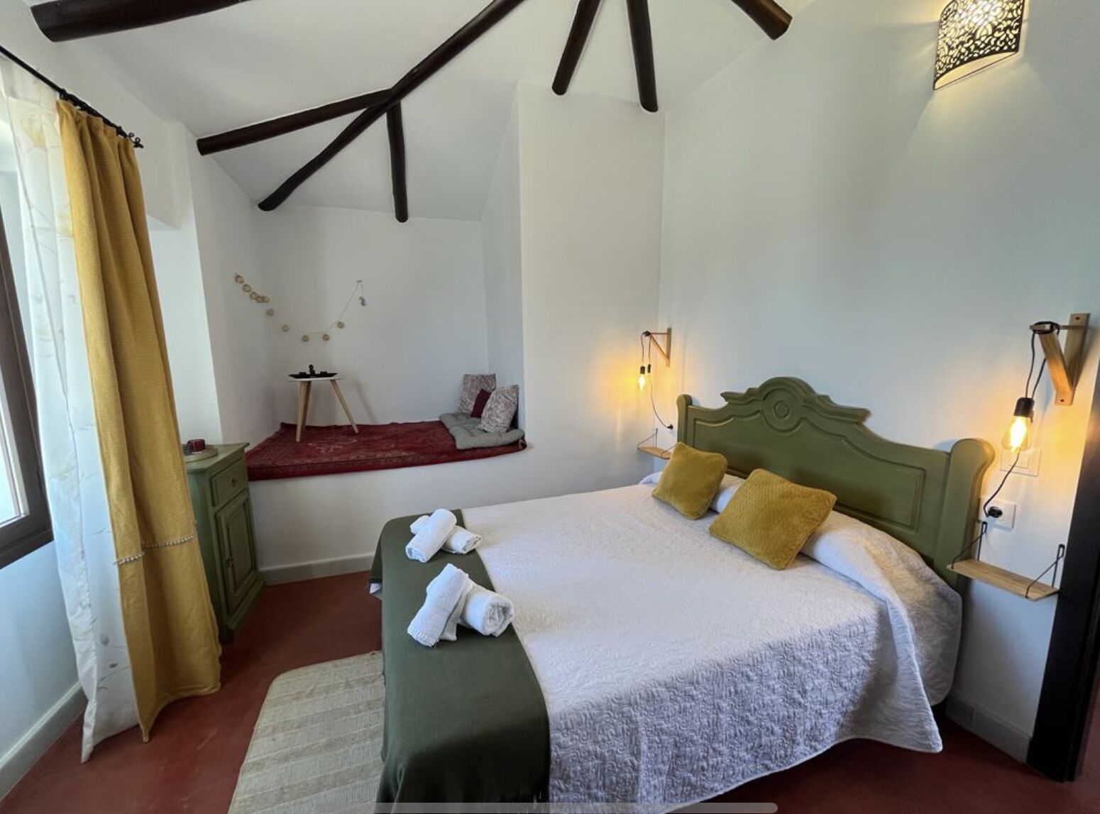 Un dormitorio con cama, lámparas de noche, una ventana con cortinas y una mesita con lámpara.