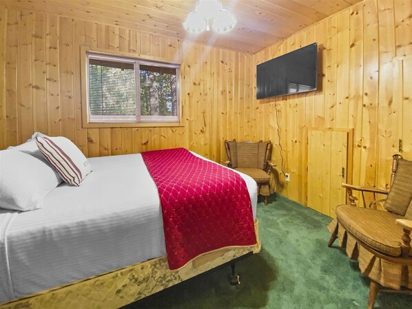 Een kamer met houten lambrisering, een bed, een stoel, een televisie en een raam.