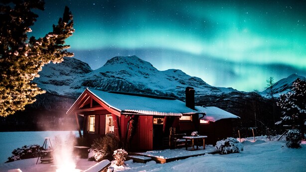A cabin with a snow-covered roof, a fire pit, and a view of the northern lights.