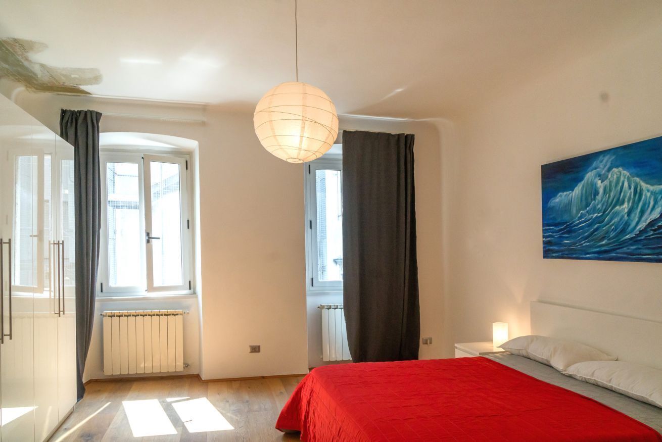 Een slaapkamer met een rode sprei, een schilderij van een golf, een hanglamp en uitzicht naar buiten door een raam.